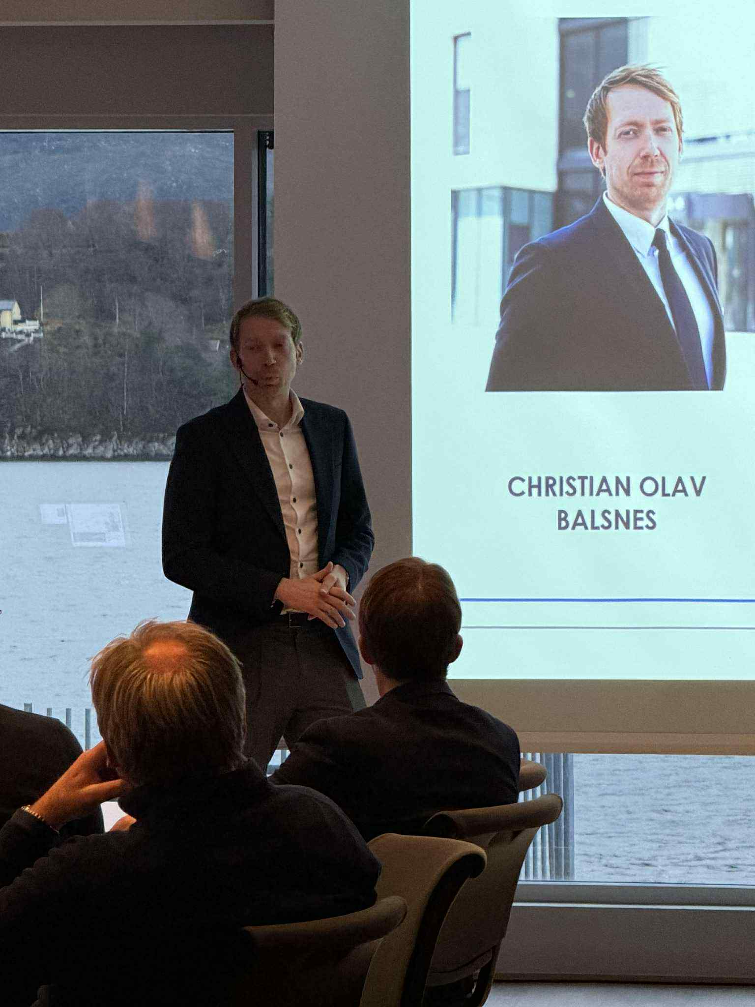 Christian Olav Balsnes med dressjakke og hvit skjorte står fremfor en forsamling. På et lerret bak står navnet hans.