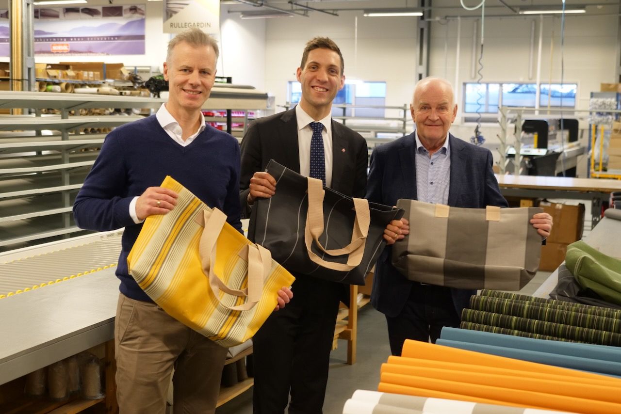 Tre personer står i produksjonslokalene hos Norsol og holder vesker laget av overskuddstekstiler fra solskjermingsproduksjonen.
