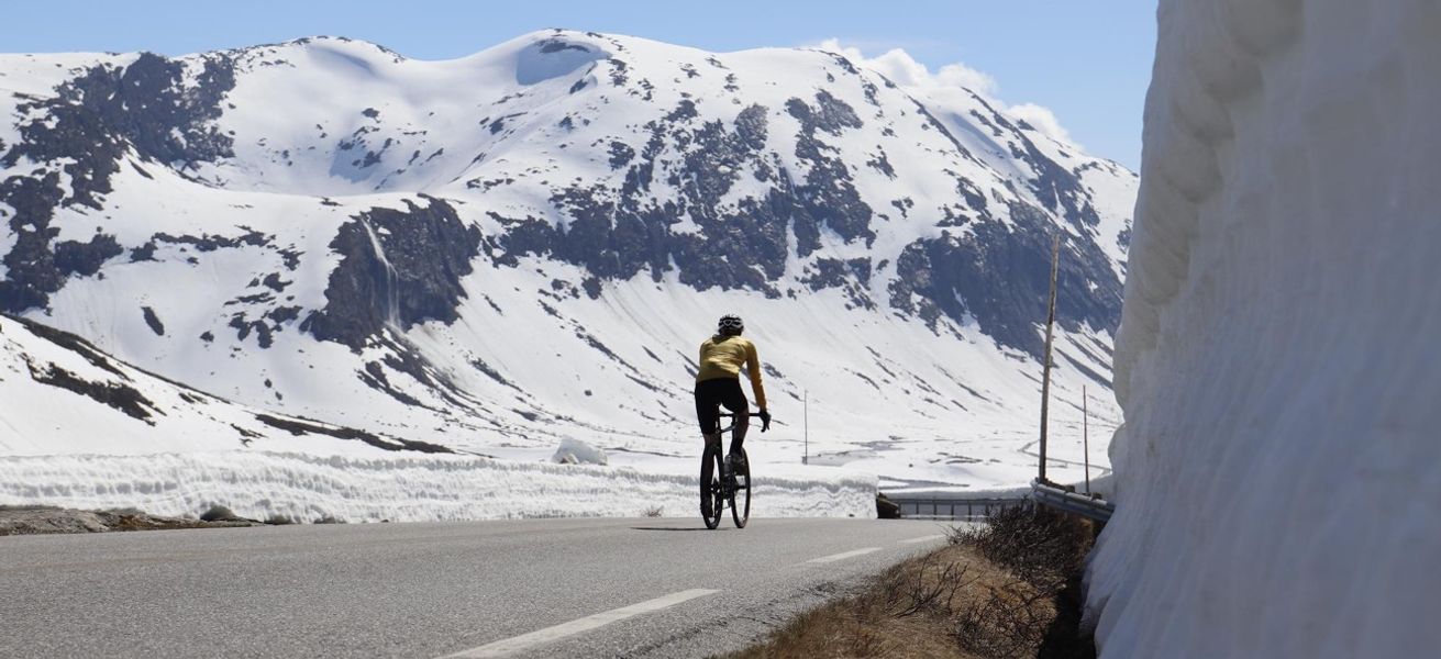 Sykkelturist på fjellvei omgitt av snø, Sunnmøre, en del av trenden Coolcation som forlenger reiselivssesongen på Nordvestlandet.