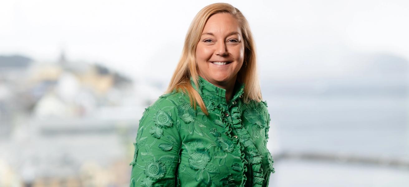 Dame i grønn bluse med lyst hår, ser i kamera og smiler. Blurret bakgrunn, utsikt mot Ålesund sentrum.