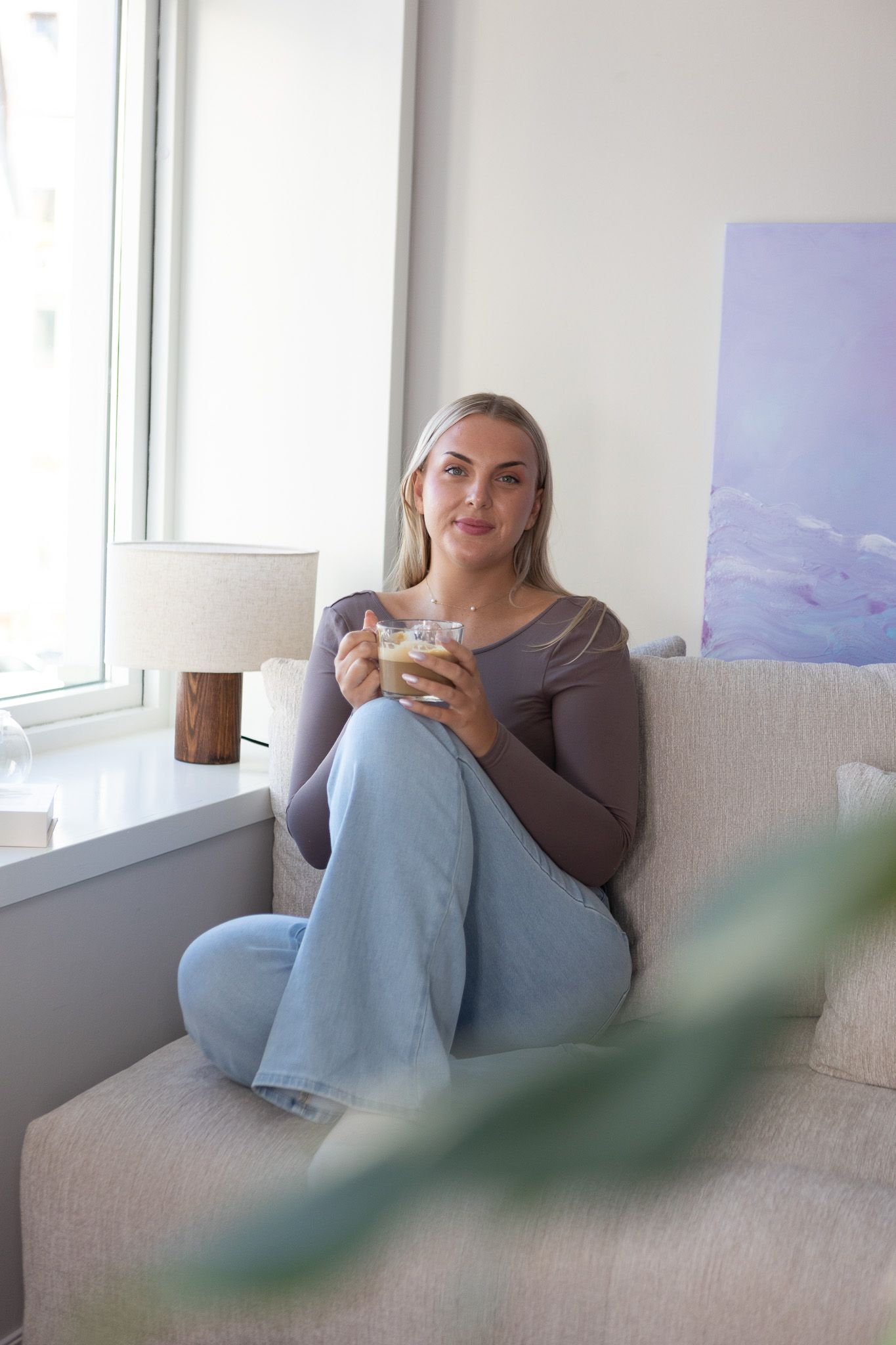 Ung kvinne i et moderne innemiljø, i sofa med en kopp kaffi. Smiler til kamera. 