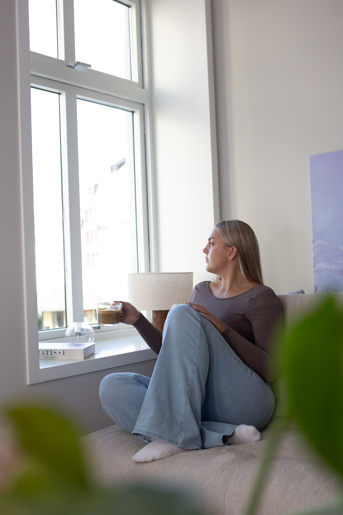 Ung kvinne som er ut av vinduet med kaffi i hånden, moderne leilighet i by. 