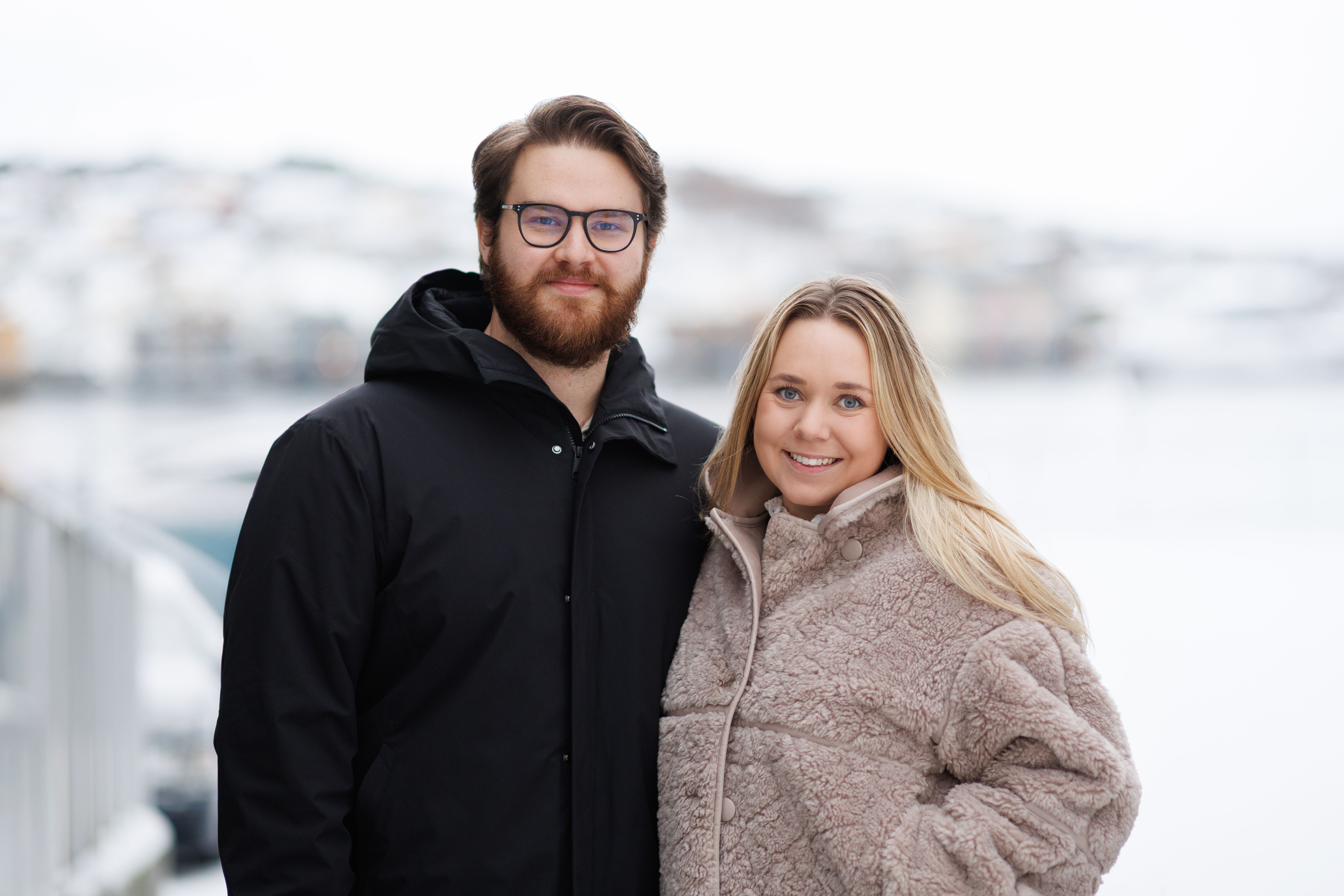 Ungt par som smiler til kamera, vinterkledd bylandskap i bakgrunnen 