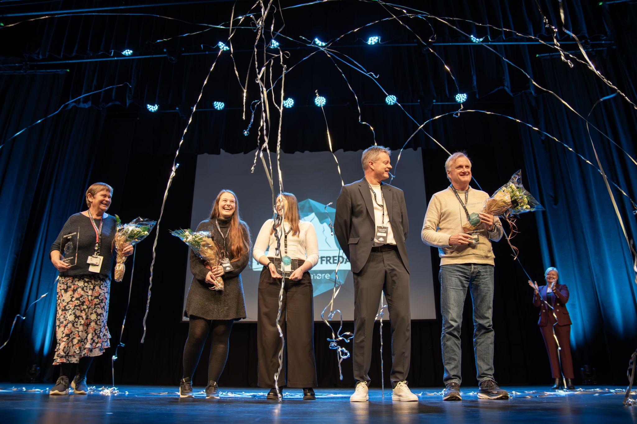 Glade mennesker på en scene, med konfettidryss. Noen holder blomster. Fabelaktig Fredag logo på skjerm i bakgrunn. 