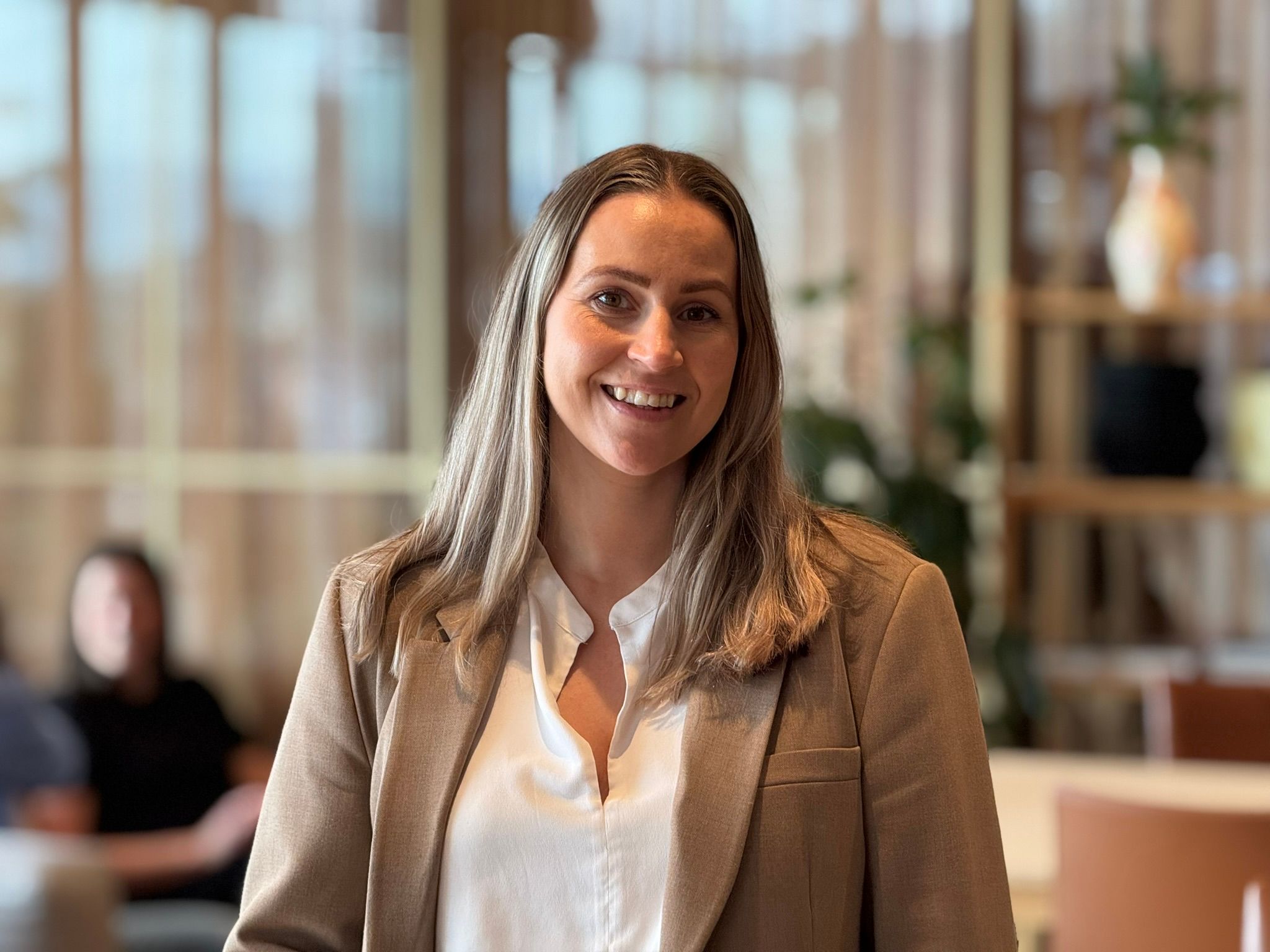 Kvinne med brunt hår, lys brun dressjakke og hvit skjorte. Ser inn i kamera og smiler vennlig.
