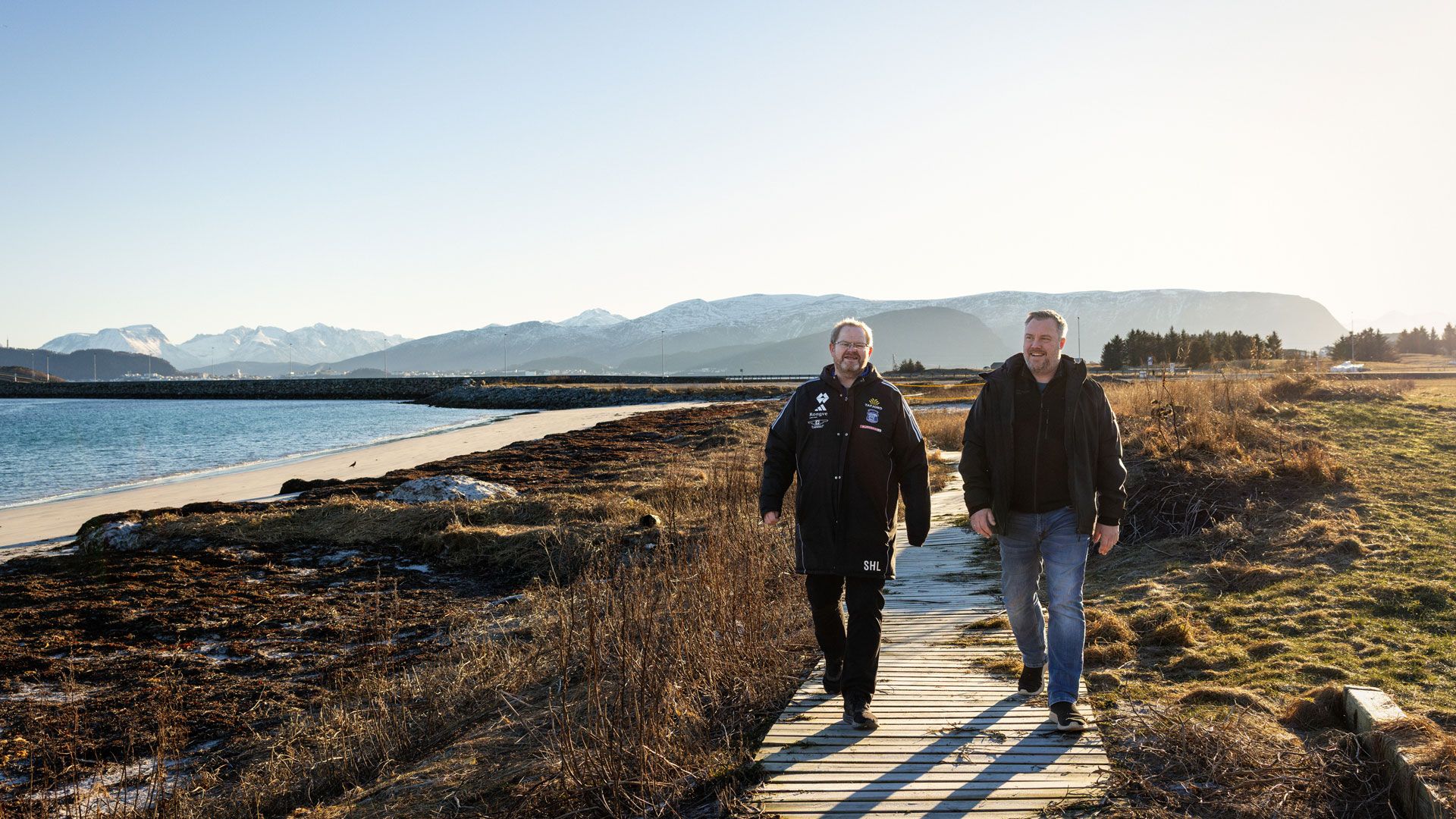 To menn går på sti ved sjøen. Det er fjell i bakgrunnen. 