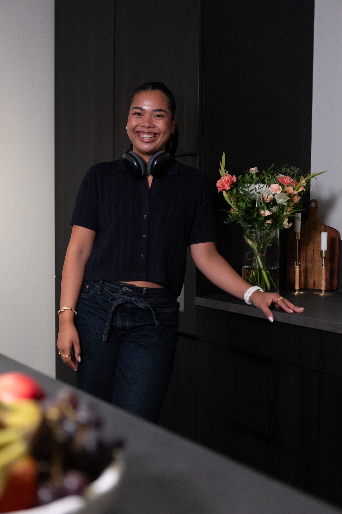 Ung kvinne med headset rundt halsen, på kjøkkenet. Hun står oppreist og smiler til kamera