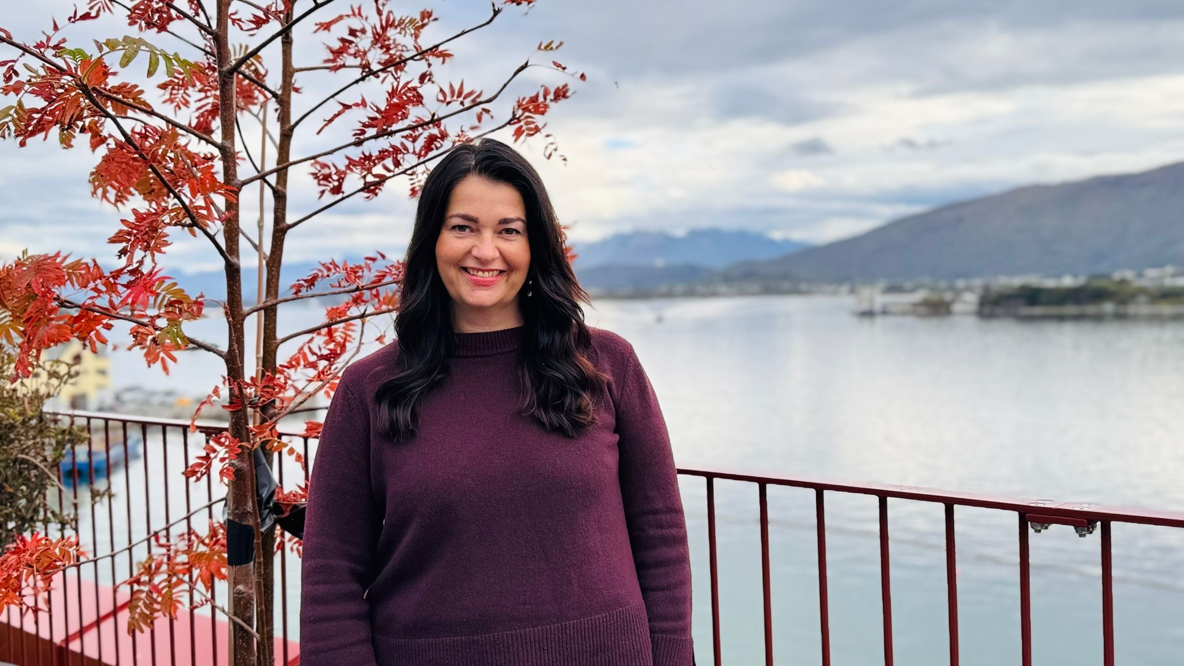 Dame med mørkt hår med fjell og fjord i bakgrunn. Ute på veranda, smiler i kamera