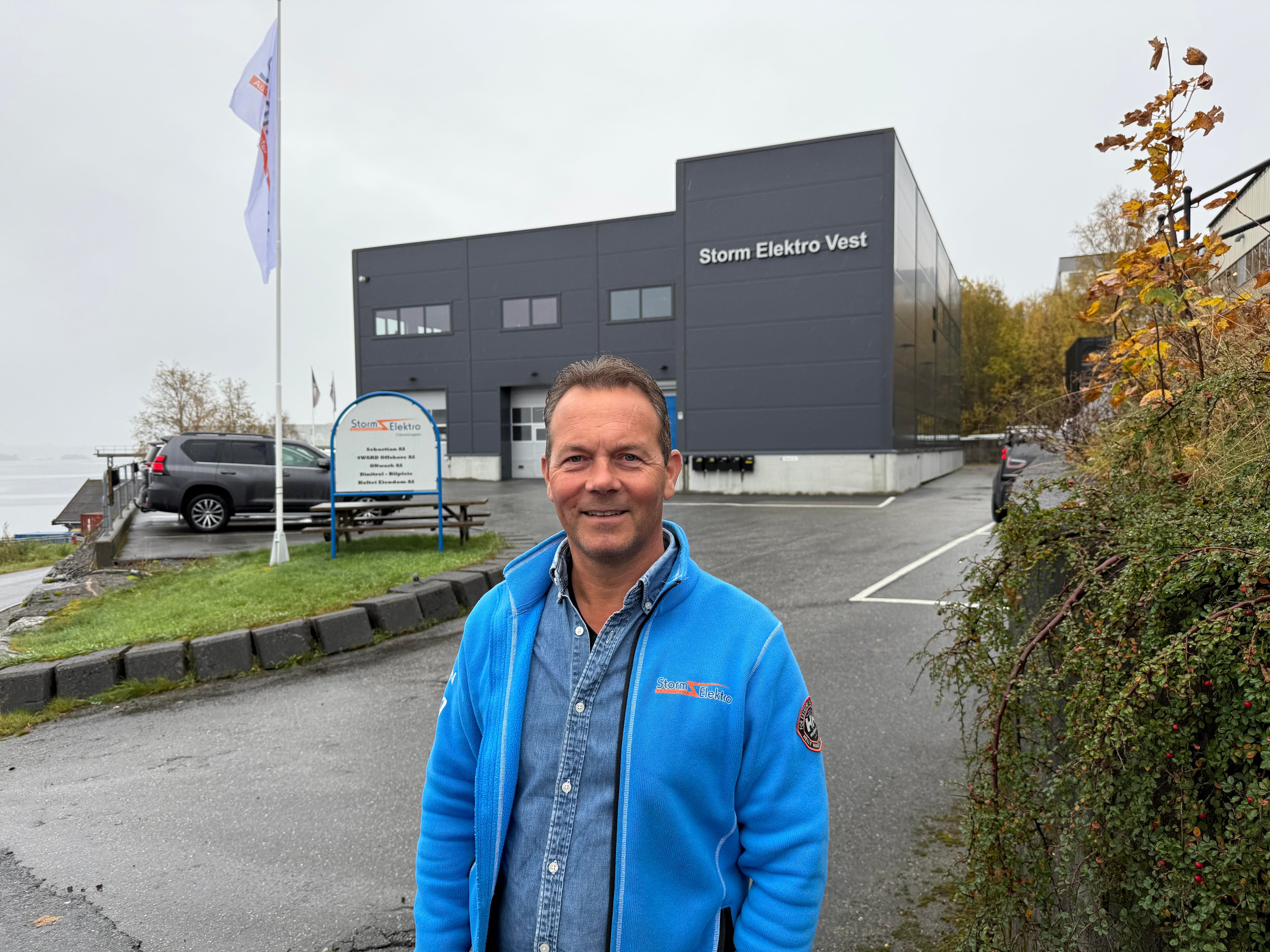 John Henry Holtet, grunnlegger av Storm Elektro smiler i kamera med Storm Elektro bygget i bakgrunnen