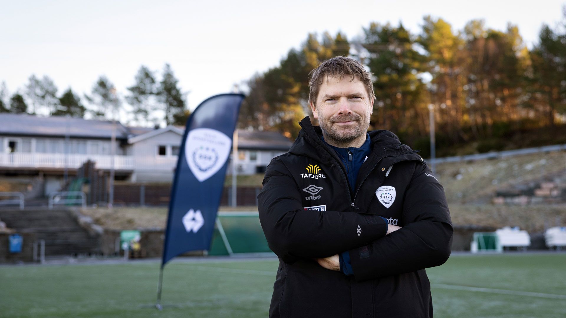 Mann står på en fotballbane, har armene i kryss og smiler